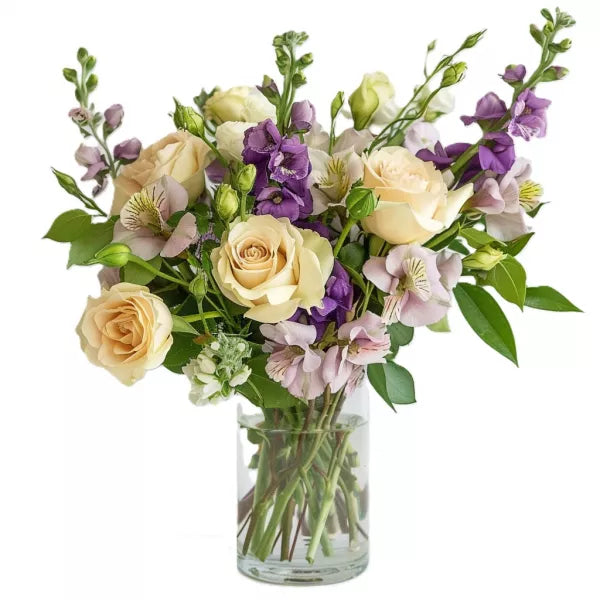 A bouquet of cream roses and purple flowers in a clear glass vase.