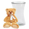 A plush teddy bear next to an empty clear glass vase.